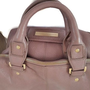 Puede incluir: Un bolso de mano de cuero malva con herrajes dorados y cierre de cremallera. El bolso tiene dos asas superiores y una correa de hombro desmontable.