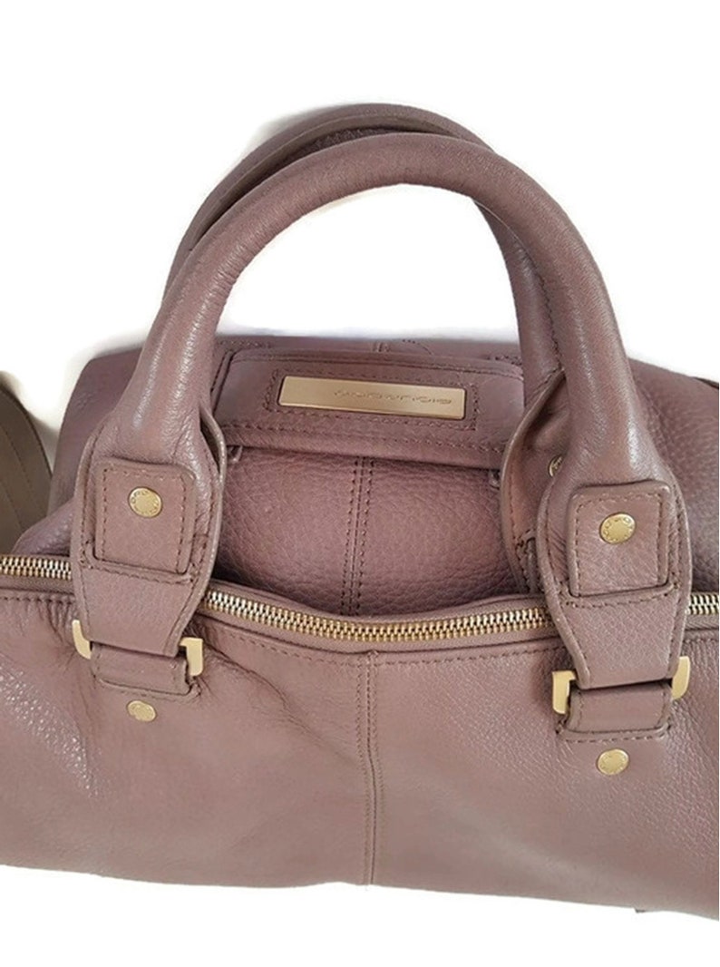 Puede incluir: Un bolso de mano de cuero malva con herrajes dorados y cierre de cremallera. El bolso tiene dos asas superiores y una correa de hombro desmontable.