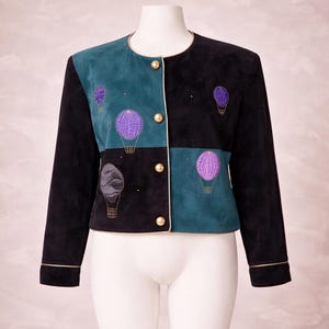 May include: A suede jacket in black and teal with a patchwork design. The jacket features embroidered purple hot air balloons and gold-colored buttons. The jacket has a round neckline and long sleeves with gold trim.