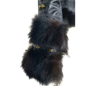 K&ouml;nnte beinhalten: Schwarze Lederjacke mit schwarzem Pelzbesatz an den &Auml;rmeln. Der Pelz ist dick und flauschig, und die Jacke hat goldfarbene Nieten an den Schultern.