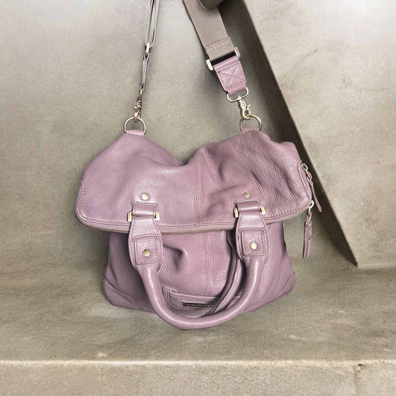 Puede incluir: Un bolso de cuero suave de color morado con una correa larga ajustable. El bolso tiene un cierre de cremallera y dos asas superiores.