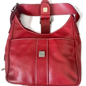 May include: A red leather shoulder bag with a curved shape and a front pocket. The bag features white stitching, a silver buckle, and a wide strap. The bag has a textured surface.