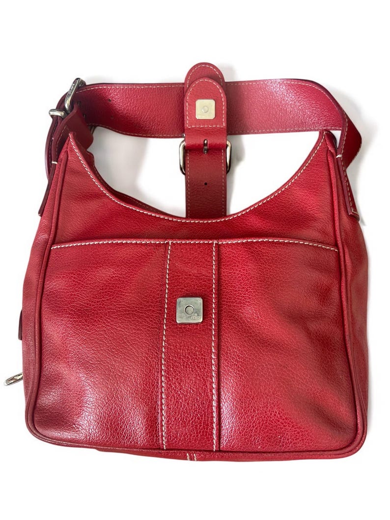 May include: A red leather shoulder bag with a curved shape and a front pocket. The bag features white stitching, a silver buckle, and a wide strap. The bag has a textured surface.