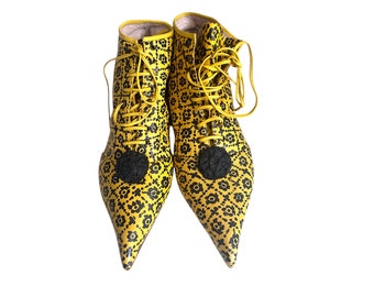 May include: A pair of yellow leather ankle boots with a black floral print and black pom-poms. The boots have laces and pointed toes.