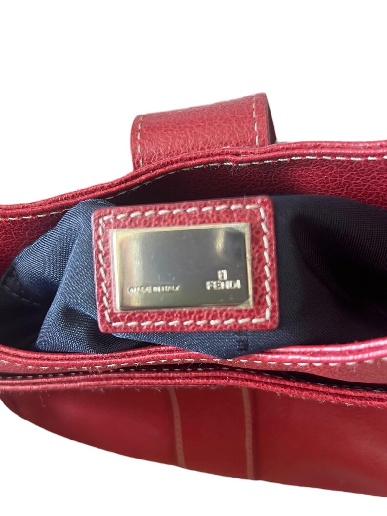 May include: Red leather handbag with white stitching. The interior is navy blue. A rectangular metal plate with the brand name "FENDI" is visible inside the bag. The words "MADE IN ITALY" are also on the plate.