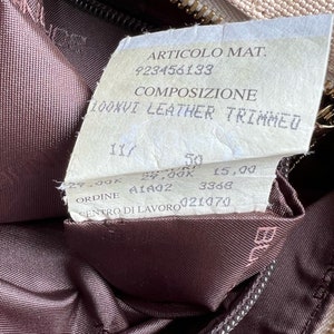 May include: A close-up of a beige leather bag with a brown leather lining. The tag attached to the bag reads "ARTICOLO MAT. 923456133 COMPOSIZIONE 100XVI LEATHER TRIMMED 29,00K 50 29,00K 15,00 A1402 3368 ORDINE CENTRO DI LAVORO 021070".