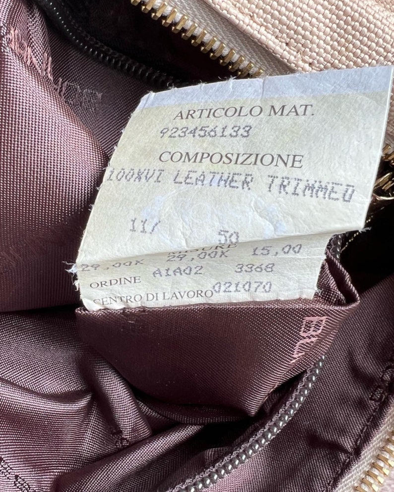 May include: A close-up of a beige leather bag with a brown leather lining. The tag attached to the bag reads "ARTICOLO MAT. 923456133 COMPOSIZIONE 100XVI LEATHER TRIMMED 29,00K 50 29,00K 15,00 A1402 3368 ORDINE CENTRO DI LAVORO 021070".