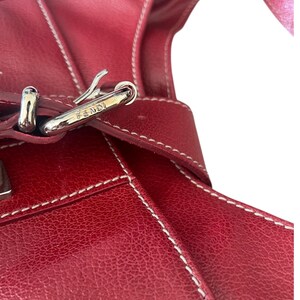 May include: Close-up of a red leather handbag with white stitching. The bag features a silver-colored buckle with the brand name "FENDI" engraved on it. The leather has a textured appearance.