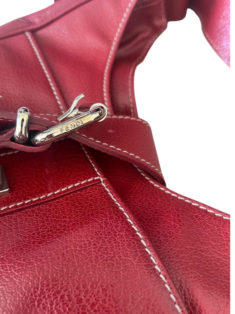 May include: Close-up of a red leather handbag with white stitching. The bag features a silver-colored buckle with the brand name "FENDI" engraved on it. The leather has a textured appearance.
