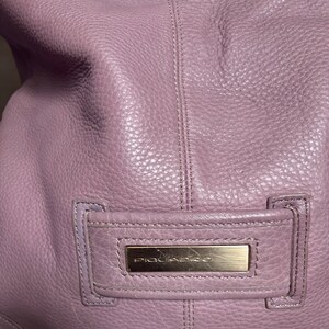 Puede incluir: Primer plano de un bolso de mano de cuero morado con una placa de metal dorado que dice "Piquadro".