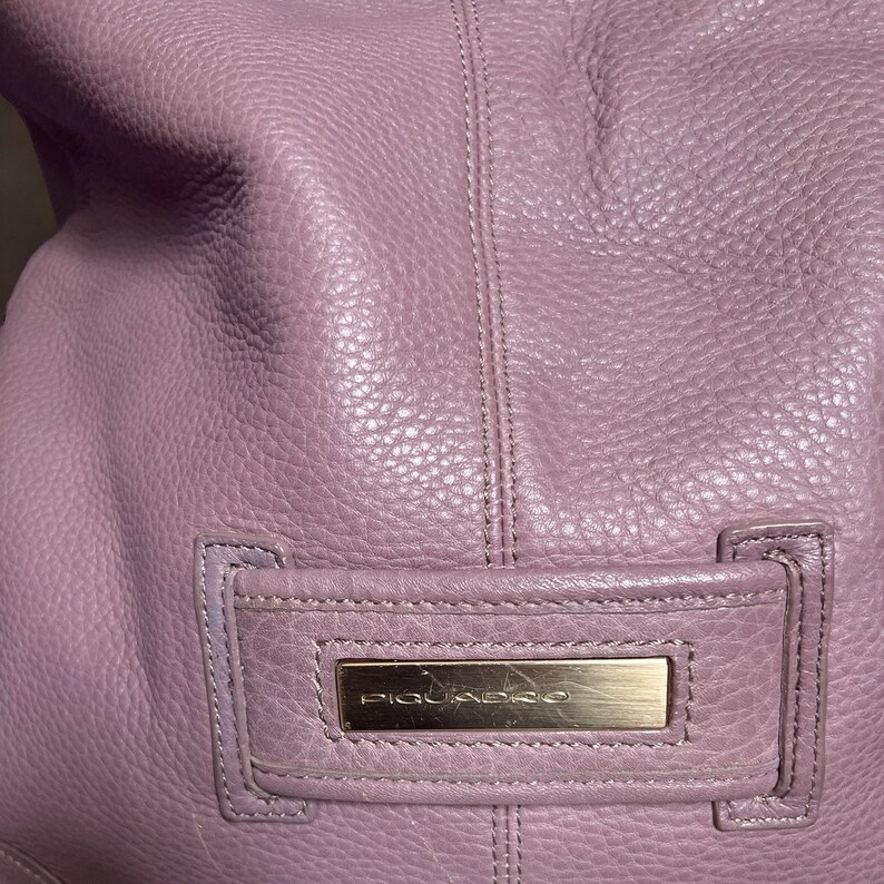 Puede incluir: Primer plano de un bolso de mano de cuero morado con una placa de metal dorado que dice "Piquadro".