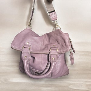 Puede incluir: Un bolso de mano de cuero de color lila claro con herrajes dorados y una correa larga ajustable.