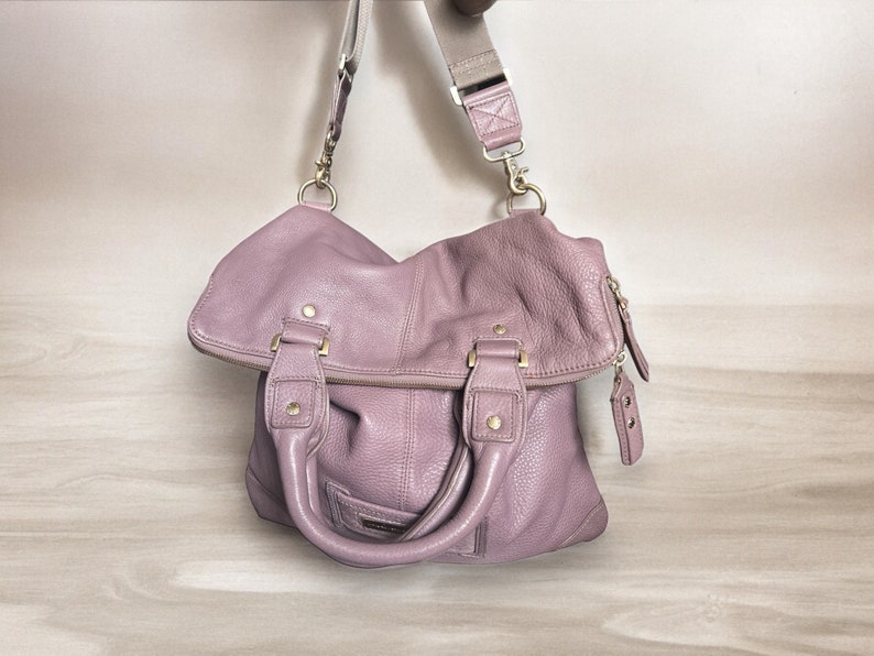 Puede incluir: Un bolso de mano de cuero de color lila claro con herrajes dorados y una correa larga ajustable.