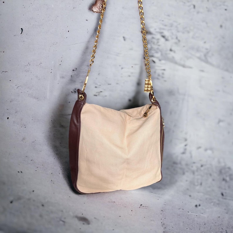 May include: A beige shoulder bag with a brown leather trim and a gold chain strap. The bag has a zipper closure and a small gold charm.