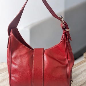 May include: A red leather shoulder bag with a single strap and silver-tone buckle. The bag features a curved shape, a flap closure with a vertical strap, and white stitching details. The bag is sitting on a wooden surface.