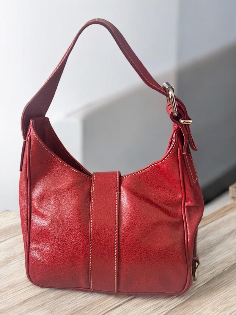 May include: A red leather shoulder bag with a single strap and silver-tone buckle. The bag features a curved shape, a flap closure with a vertical strap, and white stitching details. The bag is sitting on a wooden surface.