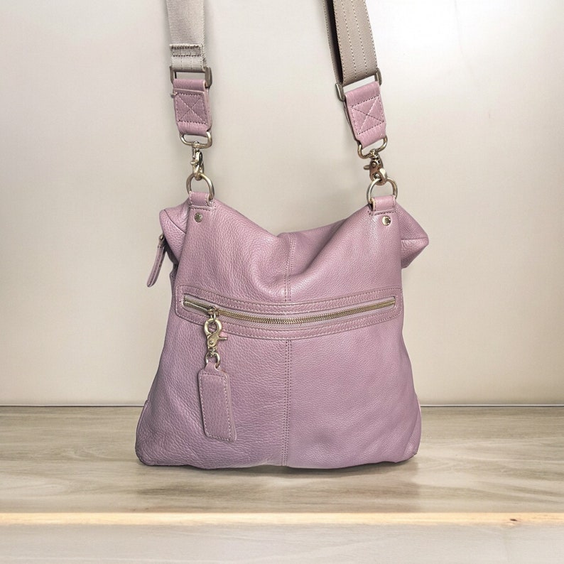 Puede incluir: Un bolso de hombro de cuero lavanda con un bolsillo con cremallera y un tirador de cremallera dorado. El bolso tiene una correa larga ajustable.