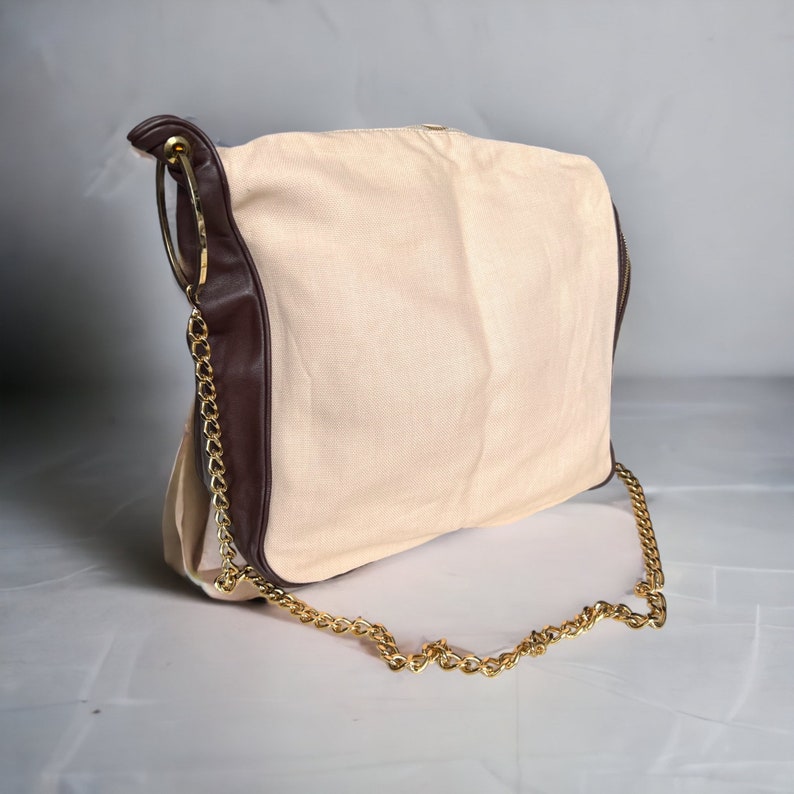 May include: A beige canvas tote bag with a brown leather trim and a gold chain strap. The bag has a large, round, gold ring at the top.