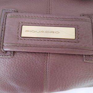 Puede incluir: Primer plano de un bolso de mano de cuero morado con una placa de metal dorado que dice "PIQUADRO".