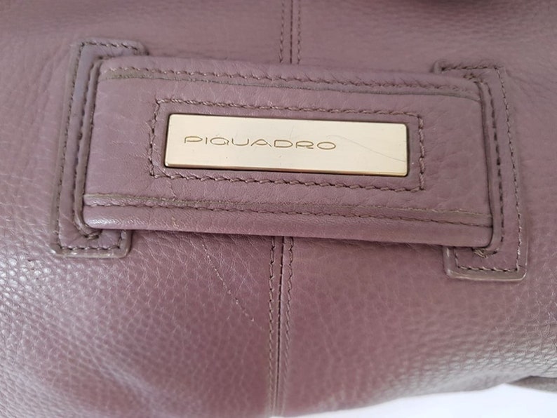Puede incluir: Primer plano de un bolso de mano de cuero morado con una placa de metal dorado que dice "PIQUADRO".