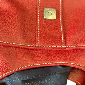 May include: Close-up of a red leather handbag with white stitching and a silver-colored square clasp. The bag has a textured surface and a dark blue interior. The text "3454/8BR148/C0J" is visible.
