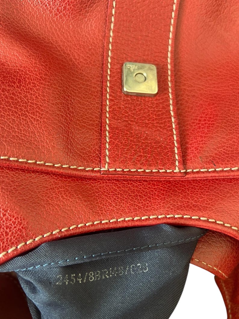 May include: Close-up of a red leather handbag with white stitching and a silver-colored square clasp. The bag has a textured surface and a dark blue interior. The text "3454/8BR148/C0J" is visible.