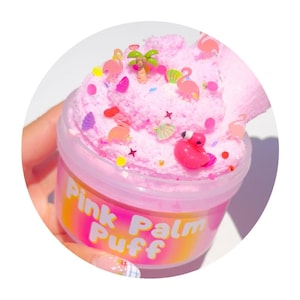 May include: A clear plastic container filled with pink slime, decorated with miniature flamingos, palm trees, and colorful confetti. The container has a pink and yellow label that reads "Pink Palm Puff".