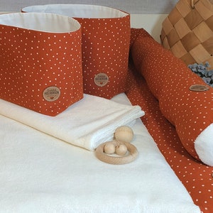 Op de afbeelding: Een witte verschoningsmat met twee oranje en witte stippen opbergmanden, twee oranje en witte stippen steun kussens, een witte handdoek en een houten bijtring.
