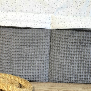 Può includere: Un organizzatore di stoffa grigio con texture a nido d'ape con due tasche. L'organizzatore ha una fodera in tessuto bianco con un piccolo motivo a pois bianchi.