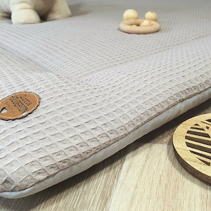 Può includere: Un tappetino da gioco in tessuto a nido d'ape beige chiaro con un'etichetta in pelle marrone che dice "Lotta  from  GERSON" e un giocattolo da dentizione in legno con tre perline di legno.