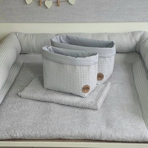 Op de afbeelding: Twee grijze opbergmanden van wafelstof met een grijze stoffen voering op een grijze verschoningsmat met grijze bumpers van wafelstof.