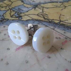 May include: A pair of white button-shaped stud earrings with four small holes. The earrings have a shiny, pearlescent finish and are set on silver posts. The earrings are displayed on a nautical map.