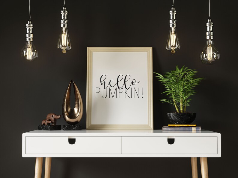 May include: A white desk with two drawers and wooden legs. A framed print with the text "hello pumpkin!" is on the desk. There are two decorative objects, a gold-colored abstract sculpture and a wooden elephant figurine, on the desk. A potted bamboo plant is on the desk. Four silver pendant lights with clear glass bulbs hang from the ceiling above the desk.