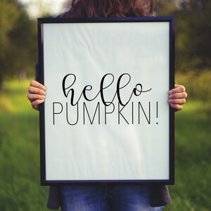 May include: A person holding a white framed sign with the text "hello pumpkin!" written in black cursive font.