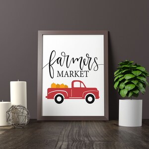 May include: A white framed print with the text "farmers market" in black cursive script. A red pickup truck with a bed full of orange pumpkins is below the text.