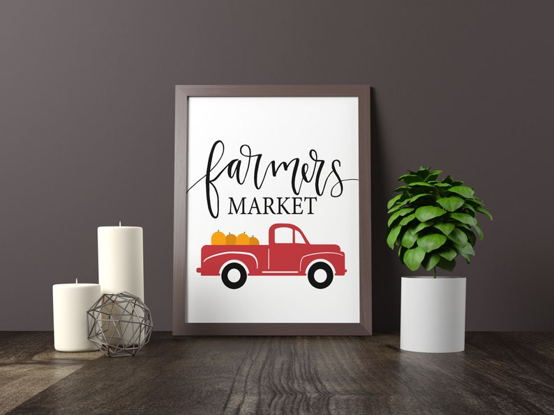 May include: A white framed print with the text "farmers market" in black cursive script. A red pickup truck with a bed full of orange pumpkins is below the text.