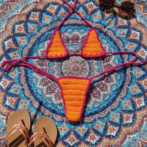 May include: An orange and pink crochet bikini set with a matching thong, laid on a blue and orange patterned beach towel. Brown leather sandals and tortoiseshell sunglasses complete the beachy scene.