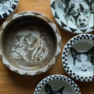 Op de afbeelding: Een verzameling keramische voerbakken voor huisdieren met handgetekende illustraties van honden en katten. De kommen hebben decoratieve randen, waaronder zwart-witte dambord- en driehoekpatronen. De kommen zijn in tinten wit, bruin en zwart.