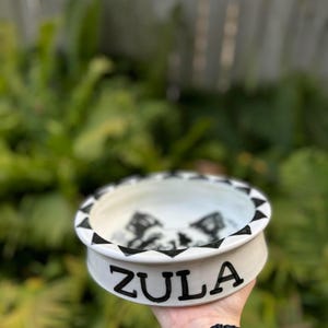 Op de afbeelding: Een witte keramische voerbak voor huisdieren met een zwart geometrisch ontwerp rond de rand en binnenkant. De naam "ZULA" staat in zwarte blokletters op de zijkant. De kom wordt tegen een vage groene achtergrond gehouden.