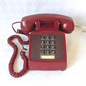 May include: A dark red landline telephone with a matching coiled cord and a handset resting on top. The base features a square keypad with twelve grey buttons, each marked with black numbers or symbols, and a rectangular label area below the keys.