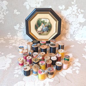May include: A collection of assorted sewing thread spools in various colors including black, white, yellow, pink, and blue, arranged in front of an octagonal framed picture featuring a painted scene of two figures in a garden setting.