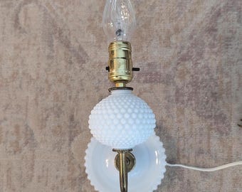 May include: A wall-mounted lamp featuring a white milk glass hobnail globe and a matching white scalloped base. The fixture includes a brass-toned metal arm and socket holding a clear, flame-shaped light bulb with a white electrical cord.