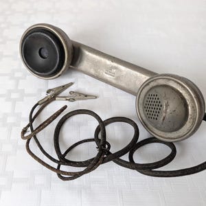 May include: Vintage silver-toned telephone handset with a black earpiece and mouthpiece. The handset has a long, rectangular body and a coiled, black cord. Two silver alligator clips are attached to the cord.