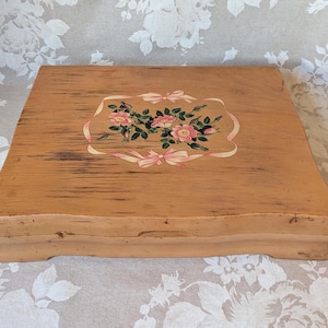 May include: A rectangular wooden box with a light tan finish and visible wood grain. The lid features a central painted design of pink roses with green leaves, framed by a decorative light pink ribbon motif. The box has a low, flat base.