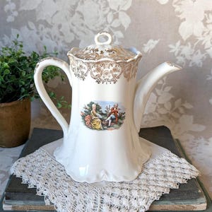 May include: Cream-colored ceramic teapot with a lid featuring gold filigree patterns. The teapot has a curved handle and spout, and a small painted scene of two figures in a garden. The teapot is sitting on a white lace doily.