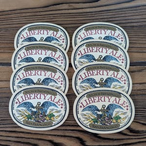 May include: Eight oval-shaped Anchor Brewing Co. Liberty Ale beer labels featuring a blue eagle perched on an anchor, wheat stalks, and hops. The text reads Liberty Ale, Anchor Brewing Co., San Francisco, and 12 fluid ounces in black and red.