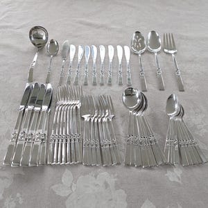 May include: A complete set of silver-colored flatware, including knives, forks, spoons, and serving pieces. The silverware is arranged on a light-colored surface, showcasing a classic design. The set includes various spoon sizes, butter knives, and forks.