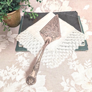 May include: A silver cake server with an ornate floral design on the blade and handle. The server rests on a white lace-edged handkerchief, atop a stack of books. A small potted plant is in the background.