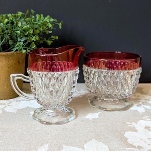 May include: A clear glass creamer and sugar bowl set. The rims are a deep red color. The glass has a diamond pattern. The creamer has a handle and a spout. The sugar bowl has a pedestal base.