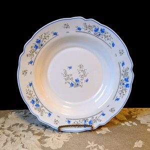 May include: A white ceramic soup bowl with a blue and gray floral pattern. The bowl has a scalloped edge and is decorated with small blue flowers and green leaves.